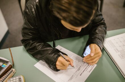 Opiskelija pöydän ääressä kirjoittaa paperiin, käden alla puhelin