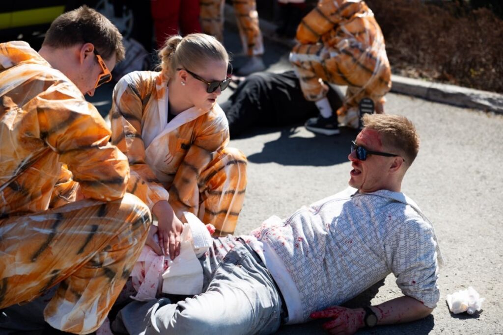 Kaksi tiikeripukuihin pukeutuntta opiskelijaa hoitamassa jalkansa loukannutta henkilöä joka makaa maassa